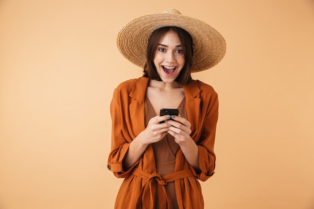 Mooie jonge vrouw met strohoed en zomeroutfit die geïsoleerd over een beige muur staat, met behulp van mobiele telefoon