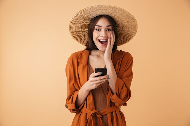 Mooie jonge vrouw met strohoed en zomeroutfit die geïsoleerd over een beige muur staat, met behulp van mobiele telefoon