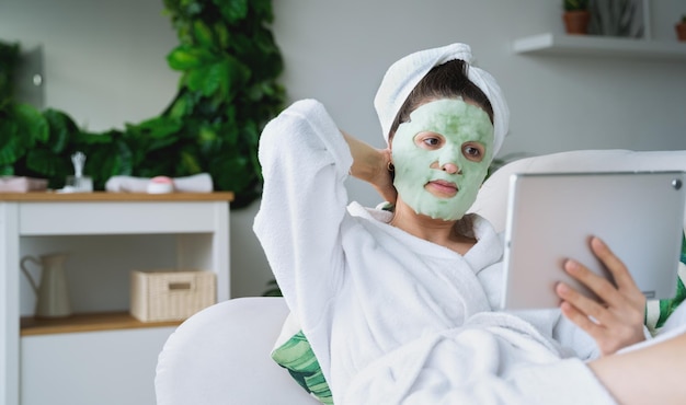 Foto mooie jonge vrouw met tabletmoisturizing groene bubbel gezichtsmaskerliggend op de bank in witte badjashanddoekself skin careanti age ochtendbehandeling voor rimpels thuisappartement met groene planten