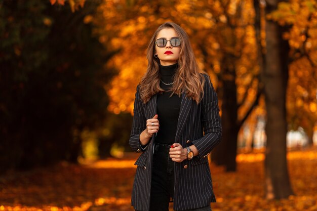 Mooie jonge vrouw met zonnebril in modieuze zakelijke kleding met een blazer en een trui loopt in het park met kleurrijke herfstbladeren. vrouwelijke zakelijke stijl en schoonheid