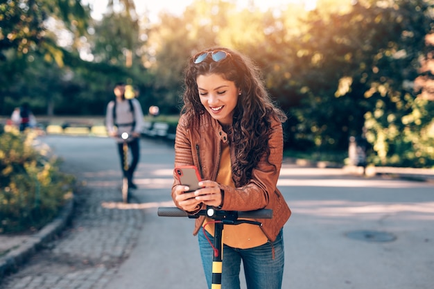 Mooie jonge vrouw op elektrische scooter kijken naar haar mobiele telefoon.