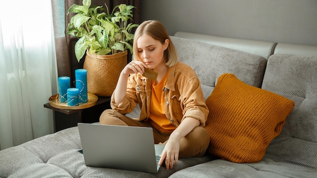 Foto mooie jonge vrouw shopper maakt online aankopen met behulp van laptop creditcards terwijl ze thuis op de bank zit vrouw doet zorgvuldig aankopen twijfelachtige attente koper lange webbanner