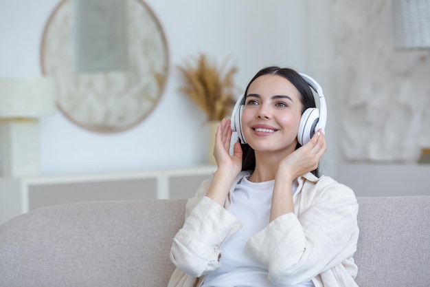 Mooie jonge vrouw thuis ontspannen luisteren naar muziek van witte koptelefoon zittend op de bank