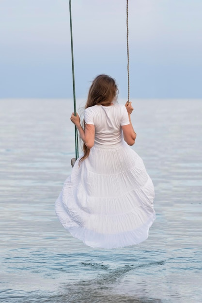 Mooie jonge vrouw zit op touw schommel op zee achtergrond Verticale frame Achteraanzicht