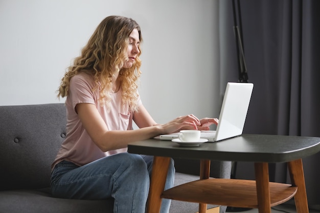 Mooie jonge vrouw zittend op de bank met een witte kop koffie die op een witte laptop werkt