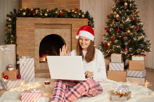 Mooie jonge vrouw zittend op de vloer met laptop op knieën, video-oproep, iemand begroeten en zwaaiende hand, geruite broek, wit overhemd en kerstman hoed dragen.