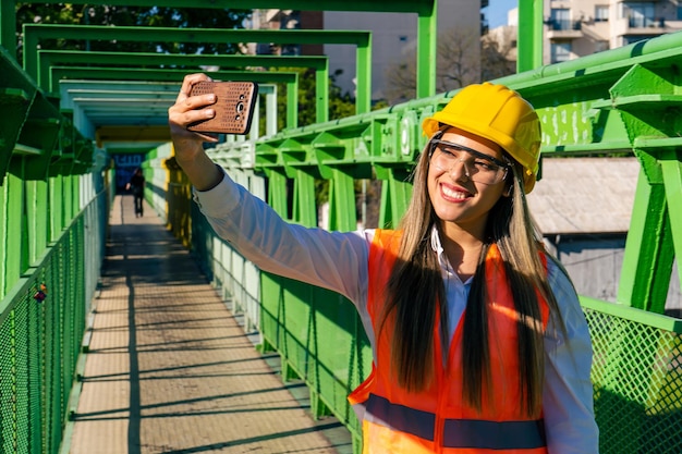 Mooie jonge vrouwelijke ingenieur of supervisor die veiligheidsuitrusting draagt en een selfie maakt met haar telefoon