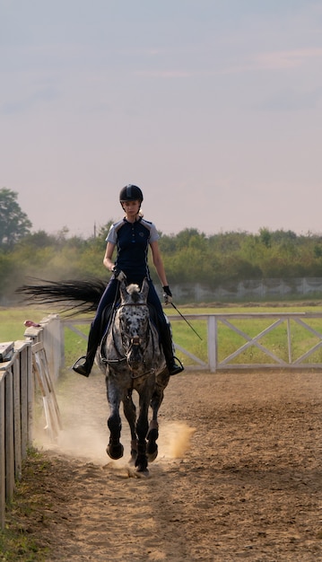 Mooie jonge vrouwelijke jockey op een paard buitenshuis. Vrouwelijke atleet rijdt een paard op open manege