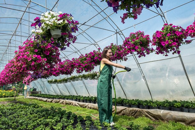 Mooie jonge vrouwelijke tuinman in schort planten in bloemenkas water geven. Concept van het verzorgen van planten