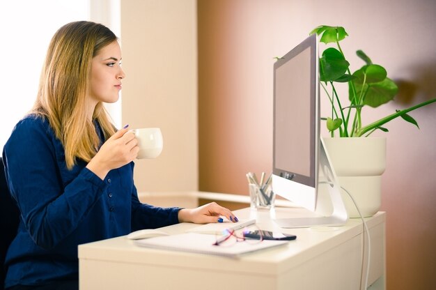 Mooie jonge zaken vrouw met behulp van computer thuis, kantoor met kopje koffie.
