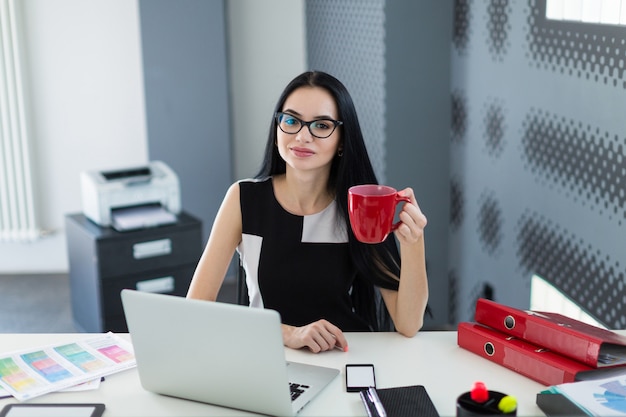 Mooie jonge zakenvrouw in zwarte jurk en bril zitten aan de tafel en werken met koffie in de hand