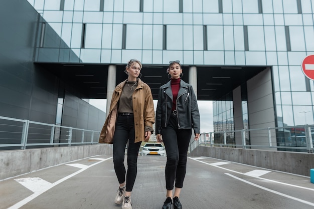 Mooie jonge zussen meisjes in een mode leren jas met stijlvolle zwarte jeans en schoenen lopen in de stad in de buurt van moderne gebouwen