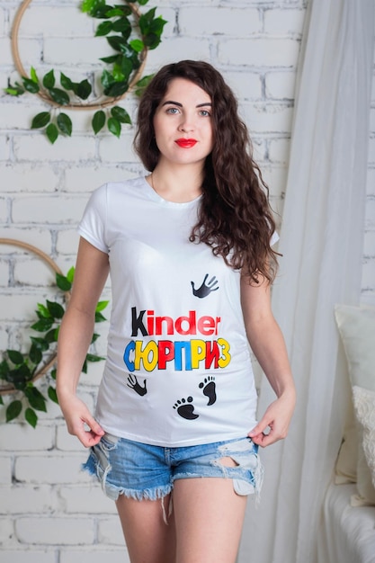 Mooie jonge zwangere vrouw met krullend haar in een t-shirt met een Kinder Surprise-print poseert in de Studio