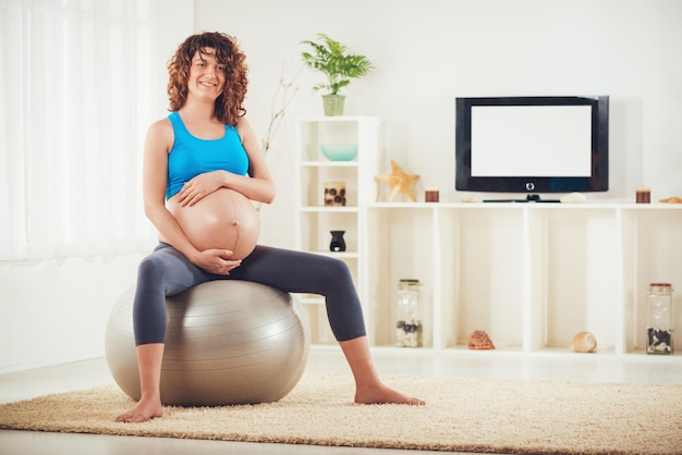 Mooie jonge zwangere vrouw zittend op een fitnessbal en thuis ontspannen.