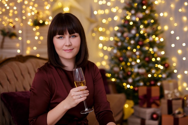 Mooie jongedame champagne drinken in ingerichte woonkamer met kerstboom en feestelijke slinger led-verlichting