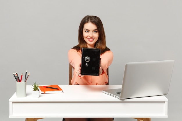 Mooie jongedame houdt metalen bank veilig voor geldaccumulatie zitten werk aan wit bureau met pc-laptop geïsoleerd op een grijze achtergrond. Prestatie zakelijke carrière levensstijl concept. Bespotten kopie ruimte.