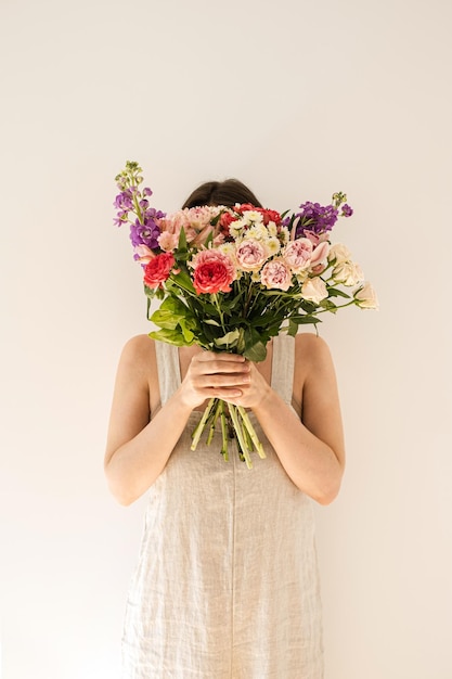 Mooie jongedame in beige gewassen linnen zomerjurk houdt boeket van kleurrijke rozen bloemen tegen witte muur