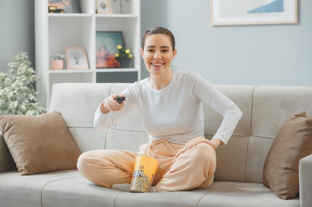 Mooie jongedame in casual kleding zittend op een bank thuis interieur met emmer popcorn houden afstandsbediening kijken naar camera gelukkig en positief glimlachend vrolijk tijd thuis doorbrengen