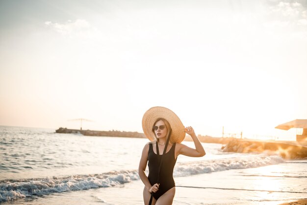 Mooie jongedame in een zwarte zwembroek en hoed met bril loopt langs het strand in Turkije bij zonsondergang. Het concept van zeerecreatie. Selectieve focus