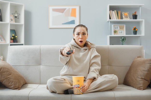 Mooie jongedame in huiskleding zittend op een bank in het interieur met een emmer popcorn die op afstand televisie kijkt, geschokt en verbaasd tijd thuis doorbrengend