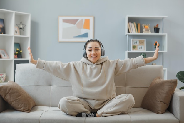 Mooie jongedame in huiskleding zittend op een bank in het interieur met een koptelefoon, blij en positief, ontspannend, zichzelf uitrekkend, oefeningen makend