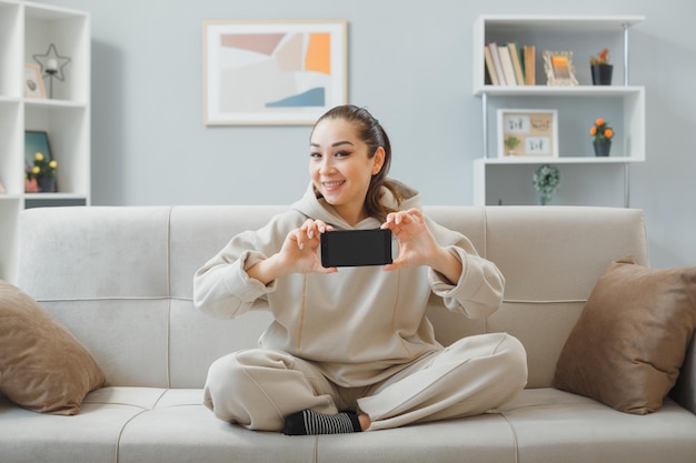 Mooie jongedame in huiskleding zittend op een bank in het interieur met smartphone die blij en positief naar de camera kijkt en vrolijk glimlacht