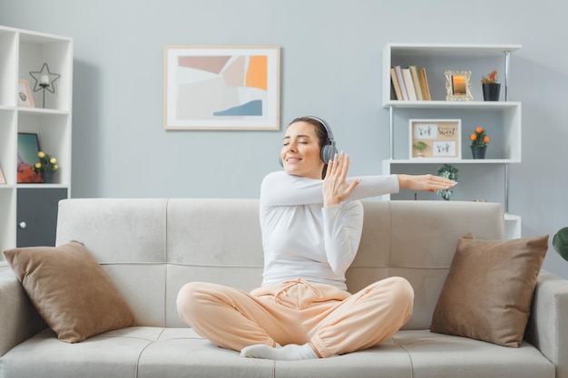 Mooie jongedame in huiskleding zittend op een bank thuis interieur met koptelefoon gelukkig en positief ontspannen zichzelf uitrekkend oefeningen makend weekend thuis doorbrengen