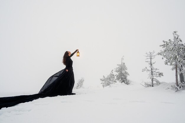 Mooie jongedame in lange zwarte jurk met oude lantaarn over winter heuvel achtergrond en sneeuwval. Sprookjemeisje op polair winterlandschap. Zwarte heks in de sneeuw.