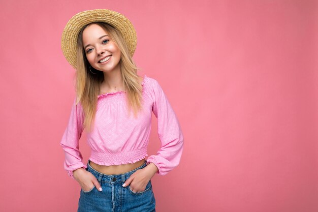 Mooie jongedame in stijlvolle roze zomerblouse en trendy strohoed. Positieve vrouw toont gezichtsemoties. Grappig model geïsoleerd op roze achtergrond met vrije ruimte.