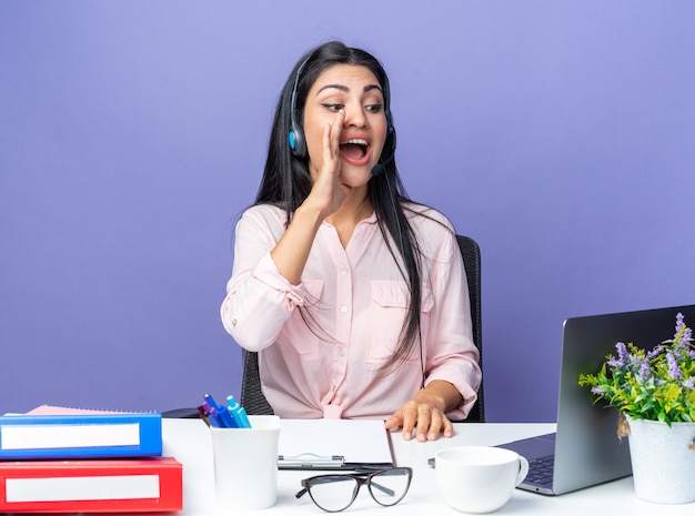 Mooie jongedame in vrijetijdskleding met een koptelefoon met microfoon die schreeuwt met de hand op de mond zittend aan de tafel met een laptop op blauw