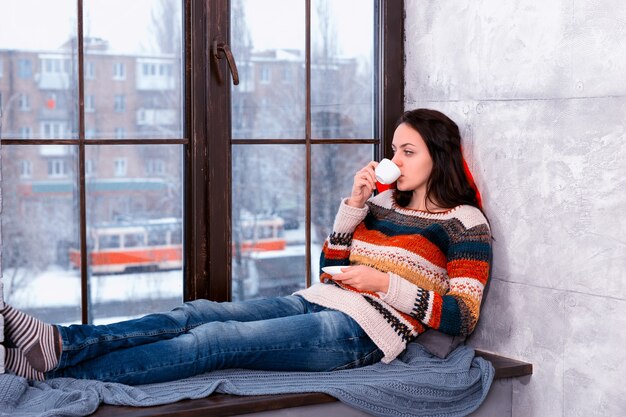 Mooie jongedame in warme gebreide trui liggend op de kussens op de vensterbank en een kopje koffie drinken