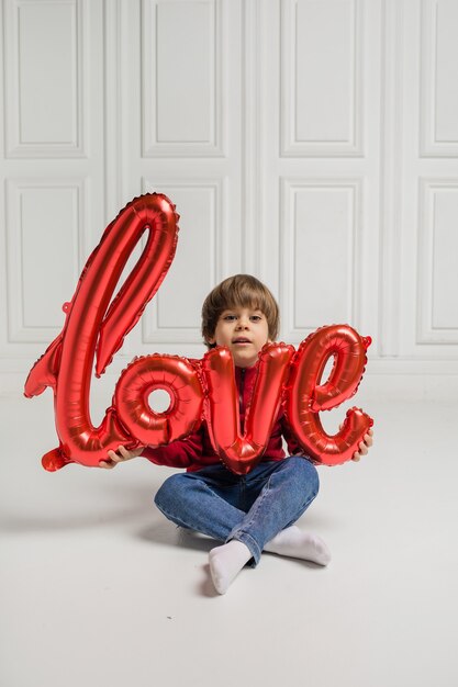 Mooie jongen zit en houdt een rode ballon op een witte achtergrond