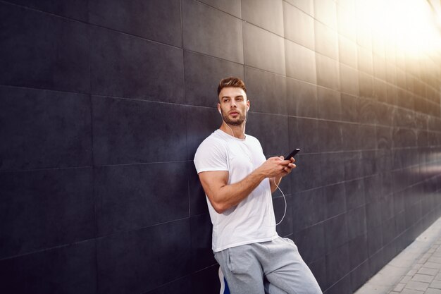 Mooie Kaukasische bebaarde blonde gespierde man in t-shirt met slimme telefoon terwijl leunend op de muur en wegkijken. In de oren zitten oortelefoons.