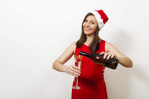 Mooie Kaukasische jonge gelukkige vrouw met charmante glimlach in rode jurk en kerstmuts champagne uit fles gieten in het glas op witte achtergrond. Santa meisje geïsoleerd. Nieuwjaar 2018 vakantie