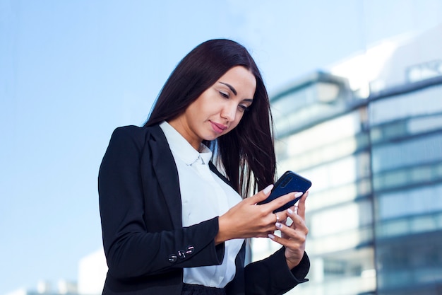 Mooie Kaukasische jonge onderneemster die haar celtelefoon bekijkt, die een bericht openlucht in een de zomerdag leest. Formeel geklede vrouw met lange nagels met behulp van smartphone buiten.