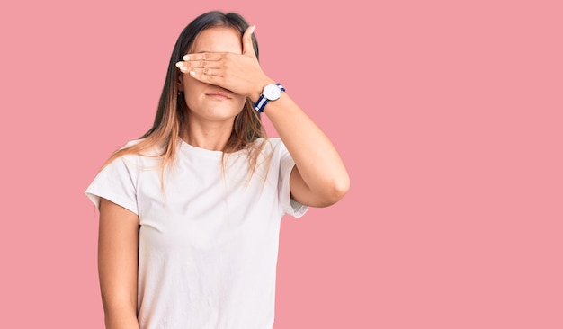 Mooie kaukasische vrouw die een casual wit t-shirt draagt dat de ogen bedekt met de hand en er serieus en verdrietig uitziet zonder te verbergen en afwijzingsconcept