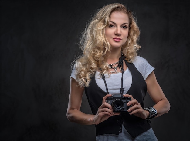 Mooie krullende blonde vrouwelijke fotograaf gekleed in een wit t-shirt en vest draagt veel accessoires en polshorloge, poseren met een camera in een studio. Geïsoleerd op donkere gestructureerde achtergrond.