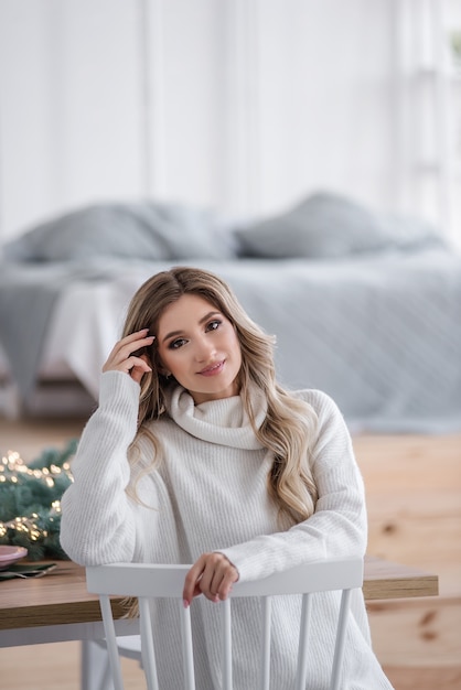 Mooie krullende blondine met lang haar bij de kerstbomen met lichtslingers. Close-up portret van een meisje met een witte glimlach in een grijze gebreide trui met een grote kraag. Kopieer ruimte, textuur