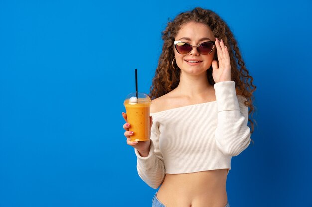 Mooie krullende vrouw die sinaasappelsap drinkt tegen blauwe achtergrond
