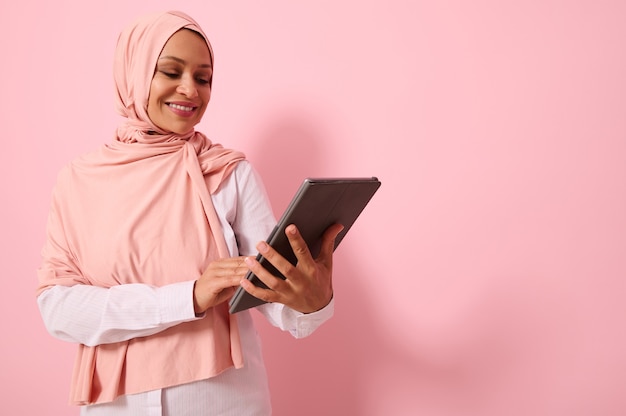 Mooie lachende Arabische moslimvrouw met bedekt hoofd in roze hijab hoofddoek, poseren tegen een gekleurde achtergrond met een digitale tablet in handen, kopieer ruimte voor tekst. Reclame geïsoleerd portret