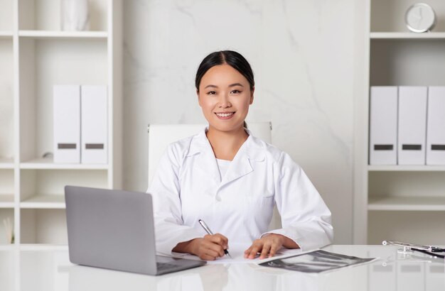 Mooie lachende Aziatische dokter dame in uniform zittend aan een bureau met laptop