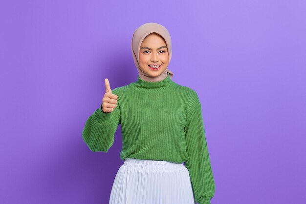 Mooie lachende Aziatische vrouw in groene trui met duimen omhoog gebaar met hand geïsoleerd over paarse achtergrond