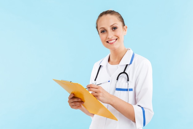 Mooie lachende blonde vrouw arts dragen uniform staande geïsoleerd over blauwe muur, het maken van aantekeningen in een notitieblok