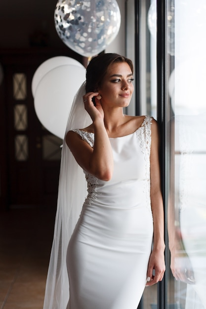 Mooie lachende bruid in mode trouwjurk binnen in de buurt van het venster. Aantrekkelijke emotionele bruid poseren in de buurt van witte ballonnen