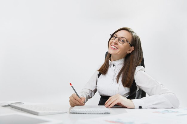 Mooie lachende bruinharige zakenvrouw in pak en bril zitten aan de balie, werken op computer met moderne monitor en documenten in licht kantoor, schrijven in notitieblok