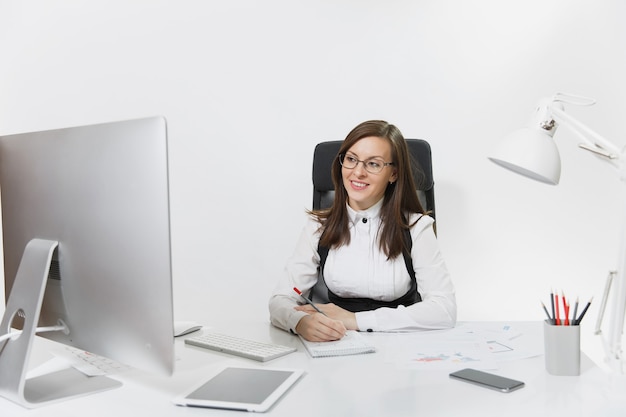 Mooie lachende bruinharige zakenvrouw in pak en bril zitten aan de balie, werken op de computer met moderne monitor met documenten in licht kantoor