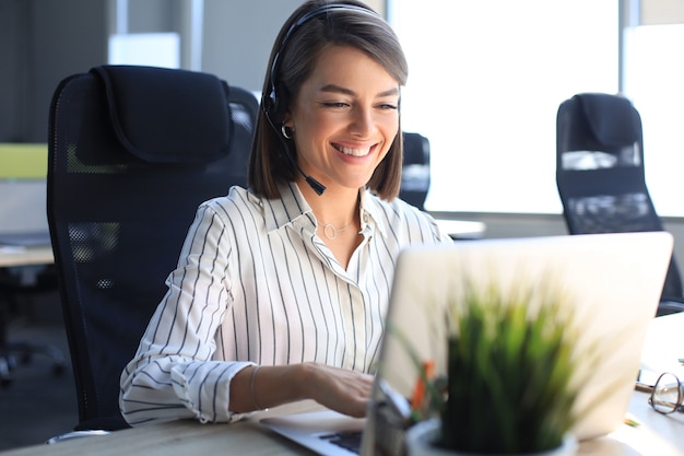 Mooie lachende callcentermedewerker in koptelefoon werkt op modern kantoor.