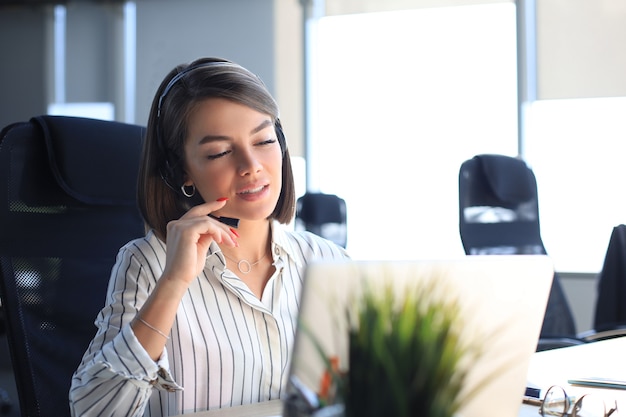 Mooie lachende callcentermedewerker in koptelefoon werkt op modern kantoor.