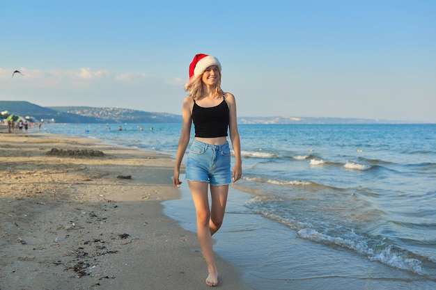 Mooie lachende gelukkig tienermeisje in Kerstman hoed wandelen langs het strand. Kerstmis en Nieuwjaar vakanties, vakanties, reizen, toerisme concept