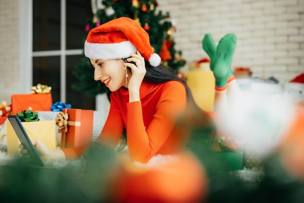 Mooie lachende gelukkige vrouw in rode jurk en het dragen van een kerstmuts ligt op de grond, omringd door kleurrijke geschenkdozen met een kerstboom op de achtergrond tablet in de hand te houden.